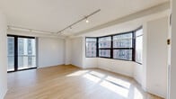 Located at 1133 North Dearborn in Chicago's prestigious Gold Coast neighborhood, this thoughtfully designed unit offers a clean, modern aesthetic throughout its single-floor layout. The spacious living area is a standout feature, boasting light hardwood flooring, track lighting, and a curved bank of large windows that frame striking views of the surrounding Chicago skyline, flooding the space with natural light. An open layout connects the living area to adjacent spaces, creating a seamless flow throughout the unit. The galley-style kitchen is well-appointed with dark wood cabinetry, white upper cabinets, stainless steel countertops, a stainless sink, and black appliances including a range, microwave, and refrigerator. The bedrooms feature plush carpeting and generously sized windows offering city views, providing a quiet retreat. The unit includes two bathrooms: one features a classic black-and-white mosaic tile floor, a walk-in shower, and a warm wood vanity, while the second bathroom mirrors the mosaic tile flooring with a sleek dark vanity, white countertop, and a large mirror accented by dark subway tile. The overall palette of whites, light woods, and dark accents gives the space a contemporary, urban character.