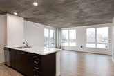 This contemporary single-floor residence showcases a striking blend of industrial and modern design elements throughout. The exposed concrete ceilings serve as a defining architectural feature, lending an urban loft aesthetic to the open, light-filled interior.

The main living and kitchen area is generously proportioned, with an open-concept layout anchored by a sleek kitchen island featuring dark cabinetry, light countertops, and a stainless steel sink. A suite of stainless steel appliances, including a dishwasher, complements the modern cabinetry. Large windows along multiple walls flood the living area with natural light and frame sweeping views of the surrounding cityscape.

The bedrooms continue the concrete ceiling aesthetic and are finished with plush carpeting for comfort. Each bedroom benefits from generous window placement, offering additional city and water views, along with baseboard heating units. Ample closet space is accessible directly from the bedrooms.

The bathrooms are clean and well-appointed, featuring light wood-look flooring, a dark vanity with a wide mirror and integrated sink, and a shower-tub combination with chrome fixtures and accessories. A well-organized hallway connects the rooms, with recessed lighting and light hardwood flooring guiding the way. Overall, the space delivers a polished, contemporary living environment with standout industrial-chic details.