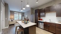 This one-bedroom, one-bathroom unit at 5350 Burnet Road in Austin's 78756 zip code offers a thoughtfully designed open-concept layout that maximizes its compact footprint. The combined kitchen and living area forms the heart of the unit, anchored by a substantial kitchen island with a quartz countertop and bar seating, ideal for casual dining or entertaining. The kitchen is outfitted with rich dark-stained cabinetry extending to the ceiling, a white subway tile backsplash, stainless steel appliances including a dishwasher, microwave, and range, and light wood-style flooring that continues throughout the main living areas. Track lighting and pendant fixtures illuminate the space effectively. The living area benefits from large windows that invite natural light and feature a ceiling fan overhead. The bedroom is a comfortable, well-proportioned room with multiple windows providing natural light and wood-style flooring. A hallway connects the bedroom to the full bathroom, which features tile flooring, dark cabinetry with a vanity, and an adjacent in-unit laundry area. A dedicated closet completes the layout. The overall palette of warm gray walls and dark wood tones creates a cohesive, modern aesthetic throughout.