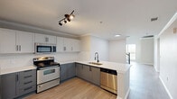This two-story, 2-bedroom, 2-bathroom residence in Dallas, TX is designed with a clean, modern aesthetic throughout. The kitchen serves as a central feature, showcasing two-tone cabinetry with white upper cabinets and gray lower cabinets, light quartz countertops, stainless steel appliances including a refrigerator, microwave, dishwasher, and electric range, and a peninsula with a sink that opens toward the living area. Light wood-tone flooring runs through the main living spaces, complementing the neutral color palette and crown molding details.

The upper level impresses with vaulted ceilings and large windows that draw in natural light and offer views of surrounding greenery. A ceiling fan adds comfort to this airy space. One bedroom features direct access to a walk-in closet equipped with wire shelving, while additional bedrooms provide generous natural light through large windows with outdoor views.

The bathroom is well-appointed with a full vanity, tub-shower combination, and tile surround. A dedicated in-unit laundry closet houses a full-size top-load washer and dryer. The 3D floor plan reveals a well-organized, functional two-story layout with a staircase connecting both levels.