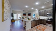This thoughtfully designed single-floor residence offers a seamless open-concept layout that connects the kitchen, dining, and living areas into one cohesive, light-filled space. The kitchen is a standout feature, boasting rich dark-toned cabinetry, granite countertops, a central island with built-in shelving, and a full suite of stainless steel appliances including a range, microwave, dishwasher, and refrigerator. Light hardwood-style flooring flows throughout the main living areas, complementing the clean white walls and recessed lighting overhead. The living room is anchored by French doors that invite natural light and provide outdoor access, paired with a ceiling fan for added comfort. A welcoming entryway with decorative accents leads into the main living space. Two well-appointed bedrooms offer plush carpeting, generous natural light through large windows, and ceiling fans. Each bedroom is served by its own bathroom; one features a tub-shower combination with granite vanity, while the other includes a tiled shower and a spacious vanity with ample storage. The overall ambiance is modern, warm, and inviting, with a cohesive neutral palette accented by contemporary fixtures throughout.