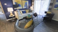 This single-floor living space showcases a thoughtfully designed interior that balances bold color choices with a comfortable, contemporary aesthetic. The living area serves as the focal point of the space, anchored by a striking navy blue accent wall that provides a dramatic backdrop for the room's decor. Two large, colorful abstract paintings hang above a neutral-toned sofa dressed with an array of decorative pillows in coordinating shades of blue, yellow, and grey.

Dark hardwood-style flooring runs throughout the space, offering a rich contrast to the lighter furnishings. A geometric-patterned area rug in navy and white defines the seating area, which is further complemented by a sleek dark coffee table and a pair of rounded grey leather accent chairs with nailhead trim detailing.

A full-length sliding glass door dominates one wall, allowing generous natural light to flow into the space and providing a direct view of the outdoors. Striped curtain panels frame the door, adding visual height to the room. The textured ceiling completes the space, contributing to its layered, well-appointed character. Overall, the space presents a cohesive and stylish living environment with a vibrant, modern personality.