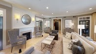 This sophisticated single-floor space showcases a refined transitional design aesthetic defined by a warm, neutral palette of taupe, greige, and crisp white. The main living area is generously proportioned, anchored by a classic fireplace with a clean white surround and a round antiqued mirror above, lending a timeless focal point to the room. Light travertine tile flooring extends throughout, reflecting the abundant natural light that streams in through multiple windows dressed in flowing gold-toned drapery panels. A standout architectural feature is the ornate wrought-iron French door that opens to a lush outdoor area, blurring the boundary between interior comfort and exterior greenery. Crown molding frames the smooth white ceilings, which are outfitted with recessed lighting that provides a soft, even illumination throughout. The seating arrangement is thoughtfully curated, combining a plush neutral sofa, upholstered wingback chairs, and charcoal accent chairs arranged around a layered area rug and nesting coffee tables. Elegant credenzas and console tables display tasteful décor and ceramic table lamps, adding warmth to the space. Additional rooms are accessible through interior doorways, further extending the functional layout of this well-designed and inviting space.
