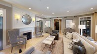This sophisticated single-floor space showcases a refined transitional design aesthetic defined by a warm, neutral palette of taupe, greige, and crisp white. The main living area is generously proportioned, anchored by a classic fireplace with a clean white surround and a round antiqued mirror above, lending a timeless focal point to the room. Light travertine tile flooring extends throughout, reflecting the abundant natural light that streams in through multiple windows dressed in flowing gold-toned drapery panels. A standout architectural feature is the ornate wrought-iron French door that opens to a lush outdoor area, blurring the boundary between interior comfort and exterior greenery. Crown molding frames the smooth white ceilings, which are outfitted with recessed lighting that provides a soft, even illumination throughout. The seating arrangement is thoughtfully curated, combining a plush neutral sofa, upholstered wingback chairs, and charcoal accent chairs arranged around a layered area rug and nesting coffee tables. Elegant credenzas and console tables display tasteful décor and ceramic table lamps, adding warmth to the space. Additional rooms are accessible through interior doorways, further extending the functional layout of this well-designed and inviting space.