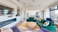 Located at 104 Washington St in downtown Dover, NH, this contemporary rooftop lounge offers a thoughtfully designed single-floor living space with an open, airy layout. The interior features warm hardwood flooring, recessed lighting, and clean white walls that create a bright and welcoming atmosphere. A built-in electric fireplace serves as a focal point along one wall, complemented by a mounted TV and built-in shelving stocked with board games and books, making it an ideal gathering space. Floor-to-ceiling windows and large black-framed glass panels line the perimeter of the room, flooding the interior with abundant natural light and framing panoramic views of the surrounding cityscape. Comfortable, colorful seating — including tufted velvet sofas and accent chairs — adds a modern, inviting character to the space. A pendant-lit dining area is visible deeper in the lounge, offering additional gathering potential. The space opens directly onto a generously sized wraparound terrace equipped with outdoor seating and dining furniture, all enclosed by sleek glass-panel railings. From the terrace, sweeping views of Dover's historic landmarks — including the iconic City Hall clock tower — provide a striking backdrop that makes this rooftop lounge a truly standout destination.