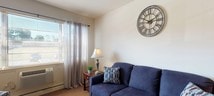 This single-floor unit at 2727 Highway 516, Old Bridge, NJ, offers a practical and comfortable layout across several well-defined living spaces. The kitchen features warm oak cabinetry, white appliances including an electric range and refrigerator, and a window dressed with light blue curtains that bring in natural light. A small dining area with a bistro-style table and chair sits adjacent, creating a cozy eat-in space.

The living area is carpeted and bright, with large windows fitted with sheer drapes allowing ample natural light to fill the room. A secondary sitting area provides additional space for relaxation, complete with its own window overlooking the outdoors and a wall-mounted television setup.

The bedroom is spacious and carpeted, featuring windows with decorative valances that allow soft natural light, along with wall-unit air conditioning. The room comfortably accommodates a full bedroom set with generous floor space remaining.

The bathroom carries a charming coastal character, with classic blue ceramic wall tiles, a tiled floor, a wood-toned vanity with a mirrored medicine cabinet, and a shower-tub combination adorned with a seashell-patterned curtain. Throughout the unit, neutral wall tones and carpeted floors create a warm, understated ambiance.
