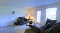 This single-floor space presents a comfortable and functional living area designed with a relaxed, residential aesthetic. The living room serves as the central gathering space, furnished with generously sized upholstered armchairs featuring decorative cushions and a classic rolled-arm design. A round glass-top coffee table anchors the seating arrangement, complemented by a small side table holding a traditional table lamp that adds warm ambient lighting to the room.

The flooring throughout is covered in plush, neutral-toned carpeting that contributes to the room's cozy atmosphere. The walls are finished in a clean, light tone, keeping the space feeling open and airy. A glass-paneled exterior door along one wall allows natural light to filter into the room, flanked by an additional window that further brightens the interior. The door provides convenient access to the outdoors while also serving as a visual connection to the exterior environment.

The overall ambiance of the space is welcoming and understated, with a layout that prioritizes comfort and practicality. Subtle decorative accents, including wall-mounted artwork and small floor accessories, add personality without overwhelming the clean, simple design of the room.