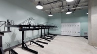 This single-floor bicycle storage facility is purpose-built to accommodate active commuters and cyclists. The room is defined by its double-tiered, black metal bike rack system, which maximizes vertical space by allowing bicycles to be stored on two levels — an efficient solution that significantly increases storage capacity without expanding the room's footprint. The lower-tier racks slide out independently, making access straightforward even in a densely packed configuration.

Along one wall, a bank of white metal lockers provides secure personal storage, offering riders a convenient place to store helmets, locks, and cycling gear. The lockers are neatly arranged in a multi-column configuration, adding both function and visual order to the space.

The room features light sage-green painted walls and a polished concrete floor, creating a clean, utilitarian aesthetic. Overhead, an exposed ceiling with industrial-style pendant lights and track-mounted fixtures delivers bright, even illumination throughout the space. The overall design is functional and well-organized, reflecting a thoughtful approach to supporting sustainable transportation options for building occupants.