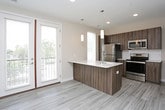 This single-floor unit presents a clean, modern aesthetic defined by light wood-look luxury vinyl plank flooring that flows through the main living and kitchen areas, transitioning to plush carpet in the bedroom. The open-concept layout connects the kitchen directly to the living space, creating a sense of openness and ease of movement.

The kitchen is a functional standout, outfitted with rich, dark wood-grain cabinetry extending to the upper walls for generous storage. A full suite of stainless steel appliances includes a refrigerator, electric range, built-in microwave, and dishwasher. Light quartz countertops offer a striking contrast to the cabinetry, and a peninsula with an undermount sink provides additional prep space and a natural division between the kitchen and living area. Recessed and pendant lighting illuminate the space effectively.

The bedroom is accessed via a sliding panel partition, offering flexibility and a quiet retreat, and features a carpeted floor and a walk-in closet with built-in shelving.

The bathroom is finished with the same wood-look flooring and includes a full soaking tub with a shower, a dual-sink vanity topped with white quartz, dark wood cabinetry with chrome hardware, and a large mirror with overhead lighting. Neutral warm-gray walls unify the entire unit with a cohesive, contemporary feel.
