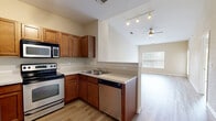 This single-floor residence offers a thoughtfully designed layout encompassing a combined kitchen and living area, one generously sized bedroom, a full bathroom, and a dedicated laundry space. The open-plan kitchen and living area forms the heart of the home, featuring warm honey-toned cabinetry, a full suite of stainless steel appliances including a refrigerator, range, microwave, and dishwasher, and a peninsula with bar seating. Light hardwood-style flooring flows throughout the main living areas, complemented by neutral warm beige walls that create a bright, welcoming ambiance. The living area benefits from a sliding glass door that opens onto a private balcony with outdoor views, and a ceiling fan adds comfort overhead. The spacious primary bedroom features vaulted ceilings, large windows fitted with blinds that invite natural light, and direct access to adjoining rooms. The bathroom is well-equipped with warm wood cabinetry, a wide vanity with a large mirror, and a combined shower and bathtub. A dedicated laundry closet houses a stacked washer and dryer unit, adding everyday convenience. Throughout, clean white trim and six-panel doors contribute to a cohesive and polished aesthetic.