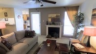 This single-story home in Amarillo, Texas, offers a warm and inviting layout designed for comfortable everyday living. The living room serves as the heart of the space, highlighted by a classic white-mantled fireplace set against a bold gray accent wall, creating a cozy focal point for the room. A ceiling fan overhead provides year-round comfort, while large windows dressed with curtains and blinds allow natural light to fill the space. Light-toned tile flooring extends throughout, contributing to a clean and cohesive aesthetic. The living area is generously sized to accommodate comfortable seating arrangements, and the open sightlines connect it seamlessly to the kitchen. The kitchen, visible from the living room, features wooden cabinetry and includes standard appliances, offering a practical and efficient workspace. A glass-panel door in the living area provides access to the exterior, enhancing the sense of openness and allowing additional natural light. The textured ceiling and neutral color palette throughout give the home a grounded, lived-in feel that balances functionality with comfort, making it a practical and welcoming residence in the heart of Amarillo.