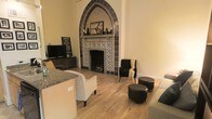 This two-floor space makes an immediate impression with its dramatic Gothic-arched fireplace, which serves as the undeniable focal point of the main living area. The fireplace is framed by elaborately patterned blue and white decorative tiles, with the pointed arch adding a distinctly historic and architectural character rarely found in residential spaces. A decorative mantle sits within the arch, adding a layered, gallery-like quality to the feature.

The living area is finished with warm-toned hardwood flooring that runs throughout, lending a natural, cohesive feel to the space. The room is furnished with a mix of upholstered seating, including a patterned accent chair, a black armchair, and rounded ottoman seating, creating a comfortable and eclectic arrangement around the fireplace. A flat-screen television mounted on a low media console completes the entertainment setup on the opposite side of the room.

The walls are painted in a soft, warm neutral tone that complements both the woodwork and the bold tilework of the fireplace. Natural light filters into the space, enhancing its inviting ambiance. The two-floor layout suggests additional living or sleeping space beyond the main level.