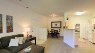 This single-floor residence offers a welcoming and functional open-concept layout that seamlessly connects the main living areas. The living room is comfortably sized, featuring neutral-toned carpeting that adds warmth underfoot, and is furnished to create a cozy, relaxed atmosphere. Soft lighting from a table lamp enhances the inviting ambiance of the space.

The dining area is positioned adjacent to the living room, providing a natural transition between the two spaces. A modest dining set sits beneath a decorative wall piece, adding a touch of personality to the area. Tile flooring in the entry and dining zone offers a practical contrast to the carpeted living space.

The kitchen is partially open to the main living area via a raised pass-through counter, allowing for easy interaction between the kitchen and the rest of the home. The kitchen features light-colored cabinetry and standard appliances, offering a clean and efficient workspace. A small decorative plant atop the upper cabinetry adds a lively finishing touch.

Throughout the space, neutral wall tones and consistent design choices create a cohesive and understated aesthetic. The layout is practical and well-suited for everyday living, with each area flowing naturally into the next.