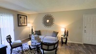 This single-floor space offers a comfortable and well-appointed living environment with a thoughtfully designed interior. The living room serves as the centerpiece, featuring rich hardwood flooring that anchors the space with warmth and elegance. Neutral-toned walls create a calm, inviting backdrop that complements the furnishings and décor. A plush sofa dressed in soft gray accent pillows sits at the heart of the room, accompanied by a sleek glass-topped coffee table and coordinating dark-framed chairs, creating a cohesive and functional seating arrangement. A bold sunburst wall mirror serves as a striking focal point above the sofa, adding visual interest and a touch of artistic flair. Framed artwork adorns the walls, contributing to the refined aesthetic of the room. Matching table lamps on either side of the seating area cast a warm, ambient glow, enhancing the cozy atmosphere. A patterned area rug ties the furniture grouping together, adding texture and depth to the overall design. The textured ceiling adds a classic architectural detail. Overall, the space presents a harmonious blend of comfort, style, and functionality, making it an ideal setting for both relaxing and entertaining.