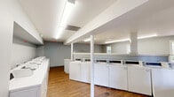 Located at 8003 SW Benaroya Lane in Huntsville, Alabama, this community laundry facility offers a well-organized and functional space designed to serve residents efficiently. The single-floor layout provides a comfortable amount of room to move freely and manage laundry with ease.

The facility is equipped with a row of commercial-grade top-load washing machines and dryers positioned along the walls, offering ample capacity for multiple users. A utility sink is conveniently integrated into the workspace, providing a dedicated area for hand-washing or pre-treating garments. A long, smooth folding table supported by hairpin legs serves as a central workstation for sorting and folding laundry, while rolling wire laundry carts are available for transporting clothes.

The interior features warm wood-look flooring throughout, complemented by light gray walls that give the space a clean and modern feel. Flush fluorescent lighting ensures bright, even illumination across the entire area. Several windows along the walls allow natural light to filter in, offering views of the surrounding greenery and outdoor areas, adding a sense of openness to the space.

A structural column and partial dividing wall define distinct zones within the room without compromising the overall flow. The space is finished with white trim and maintains a tidy, well-maintained appearance throughout.