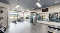 Located at 141 Nonap Rd in El Paso, TX, this fitness center offers a thoughtfully designed single-floor exercise space that provides a comprehensive workout environment. The room features durable rubberized flooring throughout, providing a safe and functional surface for a wide range of exercise activities.

The cardio zone is equipped with multiple treadmills and elliptical machines, positioned near large windows that allow natural light to fill the space and offer views of the surrounding El Paso landscape, including palm trees and nearby architecture. A decorative glass entryway door adds a stylish architectural touch to the room's entrance.

The strength training area is well-appointed, featuring a multi-functional cable machine, a full rack of dumbbells in varying weights, a weight bench, and medicine balls stored on a dedicated rack. A large wall-mounted mirror spans one side of the room, enhancing the sense of space and allowing users to monitor their form. A wall-mounted flat-screen television is positioned for visibility across the workout floor.

Additional amenities include a wall-mounted water fountain, a bulletin board for community notices, and a dedicated storage rack for accessories. Bright panel lighting installed in the dark-painted ceiling ensures consistent, even illumination throughout the space. The overall design is clean, modern, and functional, catering to a variety of fitness needs.