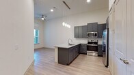 This single-floor residence showcases a thoughtfully designed open-concept layout that seamlessly connects the kitchen and living areas. The kitchen serves as the centerpiece, featuring dark gray shaker-style cabinetry that extends to the ceiling, a classic white hexagonal tile backsplash, and a granite-topped peninsula with an undermount sink—ideal for both meal preparation and casual dining. Stainless steel appliances, including a range, microwave, and refrigerator, complete the culinary space. Pendant lights hang above the peninsula, complementing the recessed lighting throughout. Light wood-style flooring flows through the main living areas, adding warmth and continuity, while a ceiling fan enhances comfort in the adjacent living space. Natural light enters through windows fitted with horizontal blinds. The bedroom offers plush carpeting, a tall window, and generous proportions. A spacious walk-in closet features multiple hanging rods and shelving for ample storage. The bathroom includes large-format tile surrounding a soaking tub and shower, a granite vanity top, and wood-look flooring, lending a clean, contemporary aesthetic throughout.