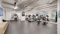 Located at 1051 Broughton Street in Vancouver's West End, this fitness facility offers a comprehensive, single-floor exercise space designed to accommodate a wide range of workout styles. The room features an exposed ceiling with industrial-style linear lighting and a clean white finish throughout, creating a bright and energizing atmosphere.

The space is thoughtfully divided into distinct functional zones. A cardio area houses multiple treadmills and stationary cycling bikes, all equipped with individual monitors. Adjacent to this is a strength training section featuring a full rack of dumbbells, barbells, weight benches, and cable machines, with floor-to-ceiling mirrors along the wall to maximize the sense of space and support proper form. A dedicated functional training area includes stability balls, balance trainers, resistance tools, and storage cubbies, along with a mounted flat-screen TV displaying workout content and instructional exercise posters across the walls. A water cooler station is conveniently positioned within the floor plan.

The durable rubber flooring provides safety and comfort underfoot, while the open layout ensures ample room to move freely between equipment. The overall design is modern, practical, and well-maintained, making it a fully equipped facility suited for both cardio-focused and strength-based training routines.