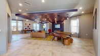 Located at 2818 Cullman Ave in Charlotte, NC, this single-floor amenity space delivers a dynamic and carefully designed environment for socializing and recreation. The layout is divided into distinct yet interconnected zones, all unified by large-format stone tile flooring that extends throughout.

The main lounge area makes an immediate impression with its deep purple ceiling accented by an array of exposed Edison bulb pendant lights that cast a warm, inviting glow. A generously sized sectional leather sofa anchors the seating area, complemented by colorful accent chairs and a bold yellow-and-gray area rug. A wall-mounted flat-screen TV and a full-size ping-pong table make this a standout entertainment hub, while curated artwork adds personality to the walls.

Adjacent to the lounge, a bright and airy social gathering area benefits from soaring ceilings and floor-to-ceiling windows that flood the space with natural light and offer views to the outdoors. This zone features a long bar-height counter with teal seating alongside a built-in booth with vibrant orange upholstery, pendant lighting, and ample room for communal gatherings.

A prep or refreshment counter with a dark stone surface, pendant lighting, and bar seating in warm orange tones provides an additional functional station. A glass-enclosed room and an elevator are visible near the entry corridor, adding convenience and accessibility to the overall space.