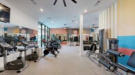 This fitness center offers a comprehensive workout environment thoughtfully designed for both cardio and strength training. The space is defined by its vibrant, contemporary aesthetic, featuring bold geometric murals in warm tones of coral, gold, blush, and teal that adorn the walls and create a visually dynamic atmosphere. Large floor-to-ceiling windows along one side of the room flood the interior with natural light while offering views to the exterior, enhancing the open and airy feel of the space. The flooring throughout is a durable speckled gym-grade surface, well-suited for high-traffic fitness use. Recessed lighting and multiple ceiling fans are installed overhead, ensuring the space remains well-lit and comfortable during workouts. The cardio section is lined with treadmills, stationary bikes, stair climbers, and cycling bikes, many equipped with personal monitors. The strength training area features a cable machine, a multi-functional weight station, a full dumbbell rack with a wide range of free weights, and medicine balls. Full-length mirrors are strategically placed throughout, amplifying the sense of space and aiding in form checks. Structural columns are integrated cleanly into the layout, and a set of double doors provides a welcoming entrance framed by a stylish herringbone-patterned accent wall.