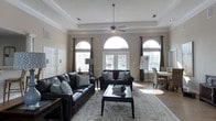 Located at 4912 Signature Dr in Myrtle Beach, SC, this single-floor space offers a refined and welcoming atmosphere defined by thoughtful architectural details and an abundance of natural light. The main living area is anchored by a coffered tray ceiling with recessed lighting and a ceiling fan, creating a sense of elevated elegance. Striking arched transom windows flank a set of French doors, flooding the room with sunlight and offering views to the exterior. Crown molding and neutral warm-toned walls contribute to a clean, classic aesthetic throughout. The living area is generously proportioned, comfortably accommodating a full leather seating arrangement centered around a decorative area rug and coffee table. Dark curtain panels frame the windows, adding contrast and a polished finishing touch. Adjacent to the main seating area, a dining space with woven chairs provides an additional area for gathering. The hard-surface flooring throughout is both durable and visually cohesive. Paneled interior doors and detailed millwork further enhance the space's traditional character. Overall, the space presents a bright, sophisticated, and functional environment well-suited to a variety of uses.