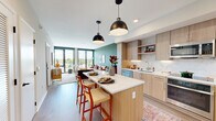 Located on the seventh floor at 3275 Washington Blvd in Arlington, VA, this 2-bedroom, 1-bathroom residence offers a thoughtfully designed open-concept layout with modern finishes throughout. Light wood flooring flows seamlessly across the entire unit, lending warmth and cohesion to every room.

The kitchen is a standout feature, outfitted with light wood flat-front cabinetry, a marble-style tile backsplash, stainless steel appliances, and a center island with a white quartz countertop and seating for two, illuminated by a pair of matte black pendant lights. The kitchen opens directly into the living area, which is anchored by a sage green accent wall and floor-to-ceiling windows that flood the space with natural light and frame sweeping views of the Arlington cityscape and treetops.

The primary bedroom continues the elevated aesthetic with large-format windows offering bright outdoor views, while the second bedroom provides additional versatile living space. A clean hallway with light wood flooring connects the bedrooms to the full bathroom, which features large-format tile flooring, a vanity with under-mount sink, and a tub-shower combination. The unit also includes in-unit utility access and ample closet storage throughout.