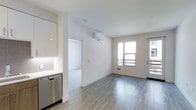 This 2-bedroom, 2-bathroom unit is designed with a clean, contemporary aesthetic throughout its single floor. The open-concept living area is anchored by light wood-style laminate flooring that extends seamlessly across the space, complementing the bright white walls and creating a cohesive, airy feel. Natural light filters in through large windows, enhancing the spacious atmosphere of the main living area. A glass-paneled door provides direct access to an outdoor space, adding a functional and inviting element to the layout. A wall-mounted mini-split HVAC unit ensures efficient climate control throughout the space. The kitchen is positioned adjacent to the living area and features stainless steel appliances, including a dishwasher, alongside white cabinetry that maintains the unit's modern, minimalist design. A doorway from the main living space leads to the additional rooms, offering a practical and well-organized floor plan. Floor vents and recessed lighting contribute to the unit's functional infrastructure. The overall design emphasizes clean lines, neutral tones, and an open feel that maximizes the sense of space.