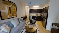 This single-floor unit showcases an open-concept design that seamlessly integrates the living, dining, and kitchen areas into one cohesive space. The living area is anchored by a light grey sofa, a round wooden coffee table, and a patterned area rug, creating a comfortable and stylish gathering space. A leather accent chair adds a bold contrast, while large wall art lends visual depth to the room.

Adjacent to the living area, a compact dining nook accommodates a small table with chairs, making efficient use of the available space. The kitchen is outfitted with rich, dark espresso cabinetry paired with light stone countertops, providing a striking contrast. Stainless steel appliances — including a refrigerator, range, microwave, and dishwasher — complete the modern kitchen setup.

Hardwood-style flooring runs throughout, contributing warmth and continuity across the space. Recessed and flush-mount ceiling lighting ensures consistent illumination throughout. The neutral wall palette and clean architectural lines give the overall space a contemporary, move-in-ready ambiance suited for modern living.