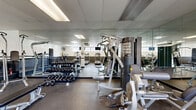 This fitness center offers a comprehensive workout environment within a single, open-plan space. The room features a drop-panel ceiling with recessed fluorescent lighting, complemented by natural light streaming through a series of horizontal windows positioned along the upper walls, providing views of surrounding greenery. The flooring throughout consists of durable, textured rubber matting, ideal for high-traffic exercise use.

The cardio zone houses multiple treadmills, an elliptical trainer, a stationary air bike, and additional aerobic equipment, each positioned beneath wall-mounted flat-screen televisions for an engaging workout experience. A ceiling-mounted fan provides additional ventilation in this area.

The strength training section is well-appointed with a multi-station cable machine, adjustable weight benches, a full rack of dumbbells ranging across various weights, and a dedicated pull-up and dip station. Full-length mirrors line the walls, visually expanding the space and allowing users to monitor their form during exercise.

Structural support columns are integrated into the layout, subtly defining workout zones without obstructing flow. A wall-mounted hand sanitizer dispenser and a padded wall accent along the lower perimeter reflect thoughtful safety considerations. The space is accessible via a dedicated entry/exit door, making this fitness center a practical and complete facility.