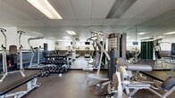 This fitness center offers a comprehensive workout environment within a single, open-plan space. The room features a drop-panel ceiling with recessed fluorescent lighting, complemented by natural light streaming through a series of horizontal windows positioned along the upper walls, providing views of surrounding greenery. The flooring throughout consists of durable, textured rubber matting, ideal for high-traffic exercise use.

The cardio zone houses multiple treadmills, an elliptical trainer, a stationary air bike, and additional aerobic equipment, each positioned beneath wall-mounted flat-screen televisions for an engaging workout experience. A ceiling-mounted fan provides additional ventilation in this area.

The strength training section is well-appointed with a multi-station cable machine, adjustable weight benches, a full rack of dumbbells ranging across various weights, and a dedicated pull-up and dip station. Full-length mirrors line the walls, visually expanding the space and allowing users to monitor their form during exercise.

Structural support columns are integrated into the layout, subtly defining workout zones without obstructing flow. A wall-mounted hand sanitizer dispenser and a padded wall accent along the lower perimeter reflect thoughtful safety considerations. The space is accessible via a dedicated entry/exit door, making this fitness center a practical and complete facility.