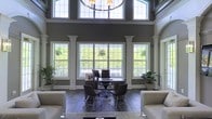 This distinguished space makes an immediate impression with its dramatically high ceilings and expansive windows that flood the interior with natural light. A row of large, grid-paned windows along the main wall frames lush greenery and outdoor views, seamlessly connecting the interior to the landscape beyond. Above, a clerestory of leaded transom windows adds an additional layer of architectural elegance while drawing even more light into the space.

The interior is defined by refined architectural details, including white decorative columns that frame the window wall, adding a classical sensibility to the overall design. Dark hardwood flooring grounds the space and contrasts beautifully with the soft gray walls and bright white trim. A statement chandelier with cylindrical shades hangs centrally, providing both ambient illumination and a focal point that draws the eye upward.

The layout accommodates a professional work area at its center, featuring a contemporary desk setup with seating for multiple individuals, making it well-suited for meetings or consultations. Flanking the workspace are comfortable lounge chairs and potted plants, contributing to a welcoming and polished atmosphere. The overall ambiance is one of sophistication and functionality, blending professional utility with an inviting, light-filled aesthetic.