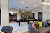 This thoughtfully designed community lounge offers a dynamic, open layout that seamlessly blends entertainment, relaxation, and social gathering spaces. The space is anchored by a full-size billiards table set atop a bold patterned area rug, with a shuffleboard table positioned nearby on decorative mosaic tile flooring — a striking design element that defines the central activity zone. Pendant lighting throughout adds warmth and a contemporary edge.

A dedicated lounge area features plush gray sofas accented with colorful throw pillows, centered around a wall-mounted flat-screen TV and a sleek linear fireplace, creating an inviting retreat for casual gatherings. Adjacent to this is a well-appointed bar counter backed by a textured geometric accent wall, illuminated by suspended linear lighting.

A standout highlight is the intimate dining nook, where a full wall of mounted vinyl records serves as a bold artistic statement above a plush, channel-tufted purple banquette. Marble-top tables paired with powder-blue accent chairs complete this retro-chic vignette, crowned by a dazzling crystal chandelier.

The space is finished with warm hardwood flooring throughout, black-framed glass double doors, and recessed lighting, combining modern design with a vibrant, social atmosphere.