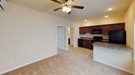 This single-floor residence offers a practical and comfortable layout with a cohesive design throughout. The open-concept living and kitchen area is anchored by plush neutral carpeting and warmed by soft, neutral-toned walls. A ceiling fan with integrated lighting serves both the living space and the adjacent kitchen, which is defined by a granite-style countertop breakfast bar that provides additional prep space and a natural division between areas. The kitchen is well-appointed with rich cherry-toned cabinetry offering ample storage, granite-style countertops, a stainless steel sink, and a full suite of black appliances including an electric range, over-the-range microwave, dishwasher, and refrigerator. Natural light filters in through a large window and a glass-paneled exterior door, lending the living area a bright and airy feel. The bedroom continues the carpeted flooring and features its own ceiling fan with light, a window with blinds for natural light and privacy, and a wall-mounted mini-split air conditioning unit for individualized climate control. The bathroom is finished with stone-pattern tile flooring, a rich wood-toned vanity, wall-mounted cabinetry for storage, and a full-size soaking tub with shower. Clean white walls and consistent trim work tie each room together for a unified, move-in-ready aesthetic.