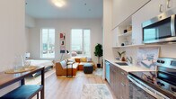 This contemporary single-floor unit offers a thoughtfully designed open-concept layout that seamlessly integrates the living, dining, and kitchen areas into one cohesive space. Impressively tall ceilings amplify the sense of openness throughout, while large windows flood the living area with abundant natural light and provide views of the surrounding streetscape.

The kitchen is fully equipped with stainless steel appliances — including a refrigerator, electric range, microwave, and dishwasher — complemented by two-tone cabinetry in white and warm wood tones, white quartz countertops, and open shelving for additional storage. A bar-height dining table near the entry offers a practical casual dining solution.

The living area features warm hardwood-style flooring that extends throughout the unit, creating a consistent and inviting aesthetic. A dedicated bedroom space provides privacy with a sliding closet door and generous room for furnishings. The full bathroom is finished with wood-toned cabinetry, a white quartz vanity, and a shower-tub combination. A separate utility area houses a stacked in-unit washer and dryer. Clean white walls, modern fixtures, and a cohesive warm-toned palette give the entire space a polished, move-in-ready feel.
