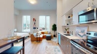 This contemporary single-floor unit offers a thoughtfully designed open-concept layout that seamlessly integrates the living, dining, and kitchen areas into one cohesive space. Impressively tall ceilings amplify the sense of openness throughout, while large windows flood the living area with abundant natural light and provide views of the surrounding streetscape.

The kitchen is fully equipped with stainless steel appliances — including a refrigerator, electric range, microwave, and dishwasher — complemented by two-tone cabinetry in white and warm wood tones, white quartz countertops, and open shelving for additional storage. A bar-height dining table near the entry offers a practical casual dining solution.

The living area features warm hardwood-style flooring that extends throughout the unit, creating a consistent and inviting aesthetic. A dedicated bedroom space provides privacy with a sliding closet door and generous room for furnishings. The full bathroom is finished with wood-toned cabinetry, a white quartz vanity, and a shower-tub combination. A separate utility area houses a stacked in-unit washer and dryer. Clean white walls, modern fixtures, and a cohesive warm-toned palette give the entire space a polished, move-in-ready feel.
