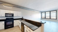 Located in Cleveland's vibrant West 9th Street corridor, this one-bedroom, one-bathroom unit offers a well-designed layout with a blend of modern finishes and industrial character. The open-concept kitchen and living area serves as the heart of the unit, featuring white shaker cabinetry, dark granite countertops, a peninsula with bar-height seating, black appliances including a range and dishwasher, a stainless steel refrigerator, and light hardwood-style flooring throughout. An exposed spiral duct runs overhead, lending an industrial loft aesthetic, while a circular pendant light anchors the kitchen space. The living area benefits from large, black-framed windows that frame sweeping views of the Cleveland cityscape and a glimpse of Lake Erie beyond. The bedroom is carpeted and offers ample space, accompanied by multiple closets for generous storage. The bathroom includes a full tub-shower combo in a clean, white finish. A dedicated laundry area adds everyday convenience. The hallway connecting the rooms features modern linear wall sconces and clean neutral tones throughout, creating a cohesive and contemporary ambiance ideal for urban living.