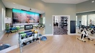 This dedicated fitness and yoga room offers a versatile, well-appointed space designed to support a wide range of workout routines. The room features light hardwood-style flooring throughout, providing a clean, durable surface ideal for both equipment-based and floor exercises. Muted sage-green walls create a calm, focused atmosphere, while large windows fitted with white blinds line multiple walls, flooding the space with abundant natural light and offering views of lush greenery outside. A full-length wall mirror expands the sense of space and allows for form-checking during workouts. The room is comprehensively equipped with a row of spin bikes for cardio training, a rack of color-coded dumbbells and kettlebells, a BOSU balance trainer, medicine balls, stability balls, and resistance equipment neatly organized on a freestanding rack. A large wall-mounted display screen with an interactive fitness platform enables guided workout sessions. A wall-mounted fold-down mat or bench adds additional versatility. An open doorway connects directly to an adjacent fitness area, making this studio part of a broader, cohesive fitness facility. Recessed lighting supplements the natural light, ensuring the space remains bright and functional at all hours.