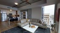This modern single-floor urban residence showcases a thoughtfully designed open-plan layout that seamlessly blends industrial aesthetics with contemporary comfort. The living area is anchored by a generously sized sectional leather sofa in a warm taupe tone, paired with a sleek gold-trimmed white coffee table and stylish decorative accents. Overhead, dark exposed ceiling tracks with industrial-style lighting fixtures add an urban edge to the space, complementing the raw concrete structural column that serves as a striking architectural focal point.

Floor-to-ceiling windows along one wall flood the living area with natural light while framing impressive elevated city views, underscoring the urban character of the residence. The open layout flows naturally from the living space toward a dining area, where dark chairs are arranged around a table, and into a well-appointed kitchen featuring white cabinetry and modern appliances. A white open shelving unit acts as a subtle divider between the living and dining zones, offering both display and storage functionality. The flooring transitions from warm wood tones in the kitchen and dining area to a deep-toned area rug in the living space, defining each zone while maintaining visual cohesion throughout.