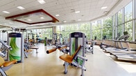 Located at 1203 Maritime Way in Ottawa, Ontario, this expansive fitness facility offers a comprehensively appointed single-floor space designed to support a wide range of fitness and wellness activities. The facility's design is defined by floor-to-ceiling windows that line the perimeter, flooding the interior with abundant natural light and offering lush, tree-lined views of the surrounding neighbourhood.

The main fitness floor is generously proportioned, accommodating an extensive array of strength training machines with modern, ergonomic designs alongside free weights and functional training equipment. A dedicated cardio zone features rows of treadmills, ellipticals, stationary bikes, and rowing machines, all positioned to take advantage of the panoramic outdoor views. A separate studio-style room with mirrored walls, a ballet barre, and open floor space provides an ideal environment for stretching, yoga, or group exercise.

A distinctive architectural focal point is a bold, floor-to-ceiling textured wood column that anchors the lounge and rest area, where stylish wooden tables and contemporary chairs offer a comfortable retreat. An elevator is integrated into this feature element. The welcoming reception area features an elegant, decorative front desk with warm wood tones and frosted glass paneling. Throughout, the space maintains a bright, professional, and motivating ambiance suited to a premium fitness environment.