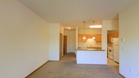 This single-floor unit features an open-concept design that seamlessly connects the living area and kitchen. The living space is finished with wall-to-wall carpet in a neutral tone, contributing to a warm and comfortable atmosphere. Clean white walls and a smooth ceiling with flush-mount lighting keep the space feeling bright and uncluttered.

The kitchen is positioned toward the rear of the open layout and is defined by a raised peninsula breakfast bar with a laminate countertop, offering both additional prep space and a natural division between the living and cooking areas. Medium-toned wood cabinetry lines the kitchen walls, providing ample storage, while pendant lights with bell-shaped shades hang above the peninsula, adding a decorative focal point. A sink is integrated into the counter beneath under-cabinet lighting, and a dishwasher is visible nearby.

A hallway extends from the main living area, leading to additional rooms. A wood-toned door is visible at the end of the corridor, suggesting access to a bedroom or utility space. The overall design prioritizes functionality and a cohesive, open flow throughout the unit.