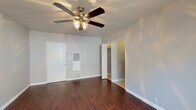 This single-floor space offers a clean, functional layout with a cohesive design throughout. Rich, dark hardwood flooring runs across the main living areas, creating a warm and inviting ambiance that contrasts beautifully with the light gray walls and crisp white trim.

The main living area is generously proportioned, bathed in natural light from a large window fitted with blinds. A ceiling fan with an integrated light fixture provides both comfort and illumination. The entry door leads directly into this open space, which also features a built-in HVAC vent panel and access to a walk-in closet enclosed by louvered bifold doors — offering ample storage potential.

The kitchen is efficiently designed, making excellent use of its footprint. White cabinetry lines the upper walls, complemented by light granite-style countertops and a stainless steel sink. The kitchen is equipped with a gas range, an over-the-range microwave, and a compact refrigerator, providing all essential cooking amenities.

The bathroom features a clean, classic aesthetic with white tile surrounding a full bathtub and shower, a pedestal sink, a framed mirror with a wall-mounted light fixture, and chrome hardware accents. Light tile flooring and neutral gray walls complete the polished look.

Overall, the space presents a practical and well-maintained interior with consistent finishes throughout.