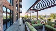 This thoughtfully designed community space combines interior comfort with outdoor living across a single level. The main lounge area is anchored by a striking floor-to-ceiling stone fireplace with a rustic wood mantel, serving as a warm focal point for relaxed gatherings. The interior features soaring ceilings, plush neutral-toned seating, and elegant multi-bulb chandeliers that cast a soft, inviting glow. Large black-framed French doors flood the space with abundant natural light while providing seamless access to the outdoor terrace. The lounge is furnished with cream sofas, wingback chairs, and accent tables, creating a refined yet approachable atmosphere. Adjacent areas include a dining nook with a wooden table and chairs, adding functional versatility to the space. The outdoor terrace extends the living experience with a generously sized composite wood deck sheltered beneath a sturdy wood-and-steel pergola. Wicker lounge furniture with vibrant green cushions and colorful accent pillows offers comfortable seating, while string lights add a welcoming ambiance. Sleek metal railings frame the terrace, revealing open views of lush treetops and the surrounding neighborhood skyline beyond.
