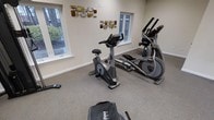 This single-floor fitness space offers a dedicated workout environment designed for comfort and functionality. The room is finished with neutral, light-toned walls and durable carpeted flooring, providing a clean and welcoming atmosphere for exercise.

The space is outfitted with a selection of modern cardio equipment, including a stationary upright bike and an elliptical cross-trainer, both from reputable fitness brands. Additional equipment such as a rowing machine and free weights can also be found within the room, offering a well-rounded range of training options.

Natural light filters in through multiple windows positioned along the walls, offering views of a lush, tree-lined outdoor setting and contributing to a bright and energizing atmosphere. Decorative wall art with a geometric, multi-toned metallic design adds a thoughtful aesthetic touch to the space, elevating it beyond a purely utilitarian setting.

The layout is open and spacious, allowing ample room to move freely between equipment. The overall ambiance is calm, tidy, and purposefully arranged, making this fitness room a practical and inviting space for a variety of workout routines.