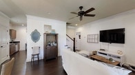This two-story home blends farmhouse charm with a clean, modern sensibility. The main living area immediately sets the tone with rich, dark hardwood floors that contrast beautifully against bright white walls and trim. A ceiling fan with a matte black finish anchors the room overhead, while recessed lighting ensures a warm, well-lit ambiance throughout. The living space is comfortably proportioned, offering room for relaxed seating arrangements alongside a dedicated media area. A dark wood bar cabinet adds a touch of rustic character and serves as a stylish focal point, accompanied by a decorative vintage-style mirror. A wooden accent chair adds to the cozy, lived-in feel of the space. The staircase, framed by white balusters and a dark wood railing, provides an elegant transition to the second floor and serves as a natural visual divider between living zones. Crown molding along the ceilings adds a refined architectural touch. The open layout encourages a smooth flow between areas, making the space feel connected and functional. Overall, the home exudes a warm, welcoming atmosphere with its carefully curated mix of rustic and contemporary elements.