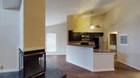 This single-floor space showcases an open-concept design that seamlessly connects the kitchen and living areas. The kitchen is outfitted with dark espresso cabinetry, granite countertops, a tile backsplash, and stainless steel appliances, including a built-in microwave and refrigerator. A peninsula with a raised granite bar provides a natural divide between the kitchen and the adjoining living space, offering additional prep or dining surface.

The living area is anchored by a corner fireplace with a dark marble surround, adding a striking focal point to the room. Vaulted ceilings rise noticeably overhead, enhancing the sense of openness and volume throughout the shared space. A ceiling fan with a pendant-style light fixture is centrally positioned, providing both function and visual interest.

Rich hardwood flooring runs continuously throughout, complementing the warm yellow-toned walls. A window in the living area allows natural light to filter in. The overall ambiance is warm and functional, with a layout that encourages easy movement and interaction between the kitchen and living spaces.