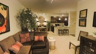 This single-floor residence showcases a thoughtfully designed open-concept layout that connects the living, dining, and kitchen areas into one cohesive and functional space. The living area is anchored by a plush, dark sectional sofa accented with vibrant throw pillows, complemented by lush indoor greenery that adds life and warmth to the room. A decorative sunburst mirror and soft accent lighting contribute to the cozy, inviting ambiance throughout.

The dining nook sits adjacent to the living space, featuring a round table with stylish chairs — an intimate setting ideal for everyday meals or entertaining guests. The kitchen is finished with rich dark cabinetry, matching black appliances including a refrigerator and microwave, and a raised breakfast bar with cushioned bar stools, providing both additional seating and a casual dining option.

Track lighting and pendant lights illuminate the space effectively, enhancing the warm tones of the neutral-colored walls and light carpeted flooring that run throughout. A textured decorative wall panel adds an artistic focal point, elevating the overall aesthetic. The open floor plan allows for excellent sightlines across all areas, making the space feel expansive, connected, and well-suited for both relaxed living and entertaining.