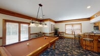This single-floor entertainment space is thoughtfully designed for recreation and socializing, combining classic craftsmanship with a warm, inviting atmosphere. Rich oak wood trim frames the walls, doorways, and windows throughout, lending a consistent and polished aesthetic to every area of the space.

At the heart of the room sits a full-size billiards table illuminated by a wrought-iron pendant light with glass shades, creating a natural focal point for gatherings. Adjacent to the pool table, a built-in wet bar with bar-height seating and an overhead microwave provides a convenient refreshment station. A dining nook nearby features a round glass-top table with bistro-style chairs, ideal for casual conversation.

A dedicated media area includes a flat-screen television, a low media cabinet, and leather recliner seating, offering a comfortable retreat for viewing. A wood-trimmed fireplace with a decorative mantel adds warmth and character to the room. Cue storage along the wall reflects the careful attention to functional detail throughout.

The space is carpeted in a bold, nature-inspired floral pattern in deep blue and tan tones, adding texture and visual interest underfoot. Multiple windows fitted with horizontal blinds allow for controlled natural light. Recessed lighting supplements the ambient illumination throughout, ensuring the space is well-lit for any activity.