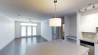 This single-floor space showcases a thoughtfully designed open-concept layout that integrates the kitchen and living areas into one cohesive environment. The kitchen is anchored by a sleek, white countertop island that flows naturally into the main living space, with a modern drum pendant light suspended above, adding a refined focal point to the area. The living room benefits from generous natural light streaming through a set of tall, shuttered French doors along the far wall, creating a bright and airy atmosphere throughout the day. A ceiling fan is centrally positioned in the living area, contributing both function and a clean aesthetic to the space. The interior is finished in a crisp, neutral white palette across the walls and ceilings, enhancing the sense of openness and light. Smooth flooring extends throughout, maintaining visual continuity between areas. A partially visible adjacent room—likely a bathroom or utility space—can be seen through an opening, suggesting a well-organized floor plan. A built-in shelf or ledge detail adds a subtle architectural element near the kitchen transition. Overall, the space presents a modern, minimalist character with practical, well-considered design choices.