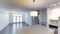 This single-floor space showcases a thoughtfully designed open-concept layout that integrates the kitchen and living areas into one cohesive environment. The kitchen is anchored by a sleek, white countertop island that flows naturally into the main living space, with a modern drum pendant light suspended above, adding a refined focal point to the area. The living room benefits from generous natural light streaming through a set of tall, shuttered French doors along the far wall, creating a bright and airy atmosphere throughout the day. A ceiling fan is centrally positioned in the living area, contributing both function and a clean aesthetic to the space. The interior is finished in a crisp, neutral white palette across the walls and ceilings, enhancing the sense of openness and light. Smooth flooring extends throughout, maintaining visual continuity between areas. A partially visible adjacent room—likely a bathroom or utility space—can be seen through an opening, suggesting a well-organized floor plan. A built-in shelf or ledge detail adds a subtle architectural element near the kitchen transition. Overall, the space presents a modern, minimalist character with practical, well-considered design choices.