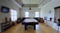 This single-floor amenity space is designed as a social and recreation lounge, offering a well-appointed environment for leisure and gathering. At the heart of the room stands a full-size billiards table, finished in dark wood with a light felt surface, and illuminated by a striking tiered pendant light that hangs from a coffered tray ceiling above. The tray ceiling itself is a notable architectural feature, adding depth and a sense of refinement to the space. Light hardwood flooring extends throughout, lending warmth and a clean, contemporary feel. Natural light pours in through multiple windows fitted with horizontal blinds, keeping the atmosphere bright and open. The perimeter of the room is furnished with dark, cushioned sectional sofas and loveseats, providing ample seating for guests to relax and socialize. A pair of bar-height stools and side tables complete the lounge setup, offering additional casual seating options. The overall ambiance is modern yet welcoming, striking a balance between a recreational game room and a comfortable communal lounge. Crown molding along the upper walls adds a polished, finished look to the space, reinforcing its well-designed character.